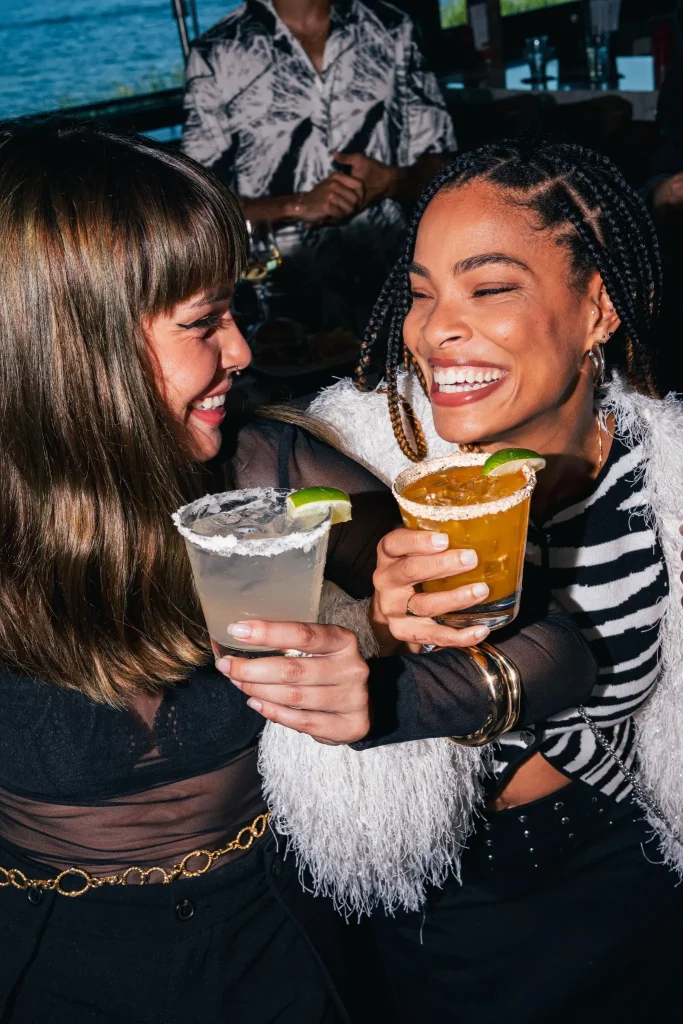 Two women smiling and laughing as they clink cocktail glasses, each garnished with a lime wedge and a salt rim, in a lively social setting.