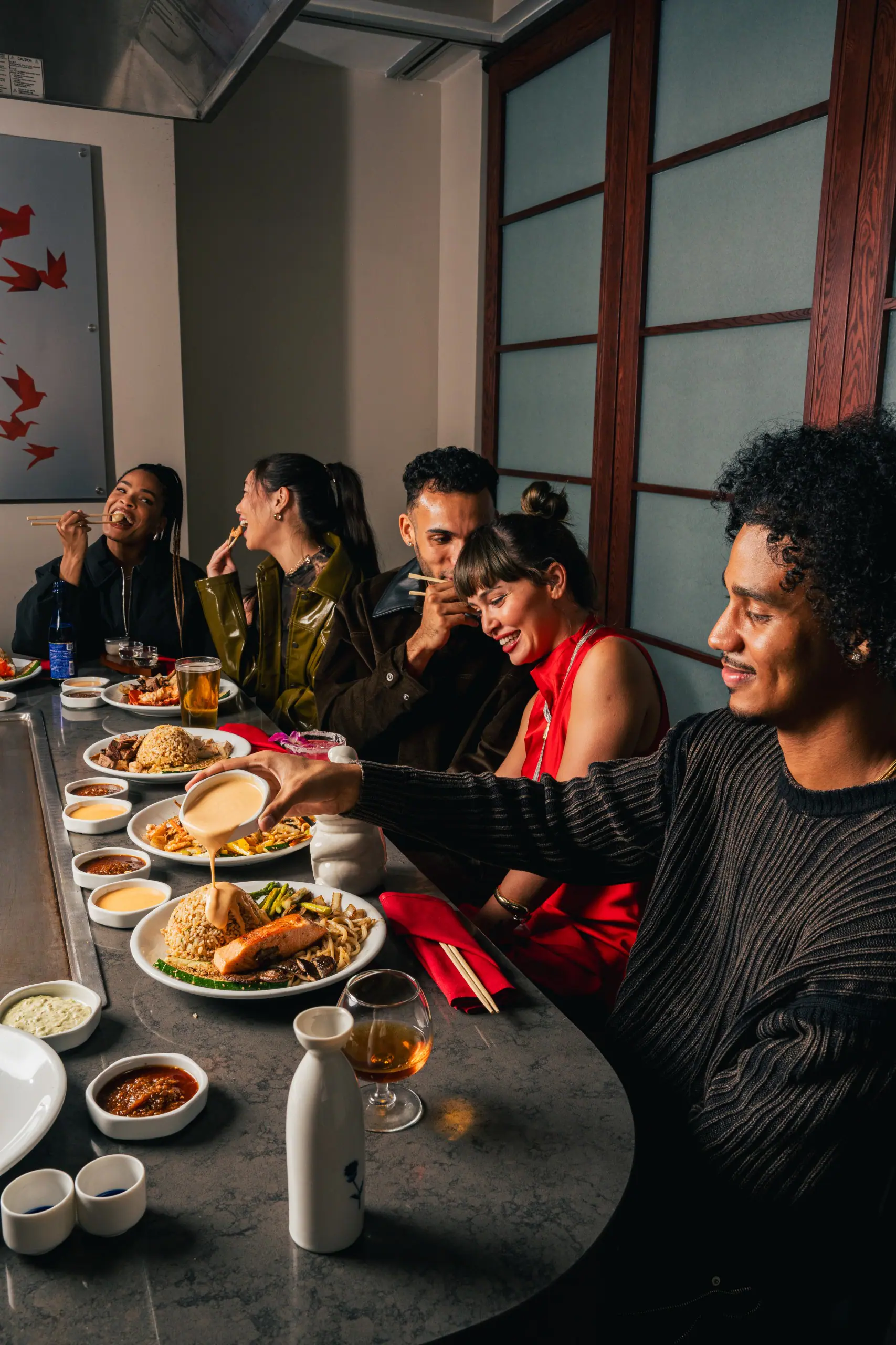 A group of five friends seated dining. Plates of hibachi fried rice, grilled salmon, vegetables, dipping sauces, and drinks. One person pours sauce over their meal.