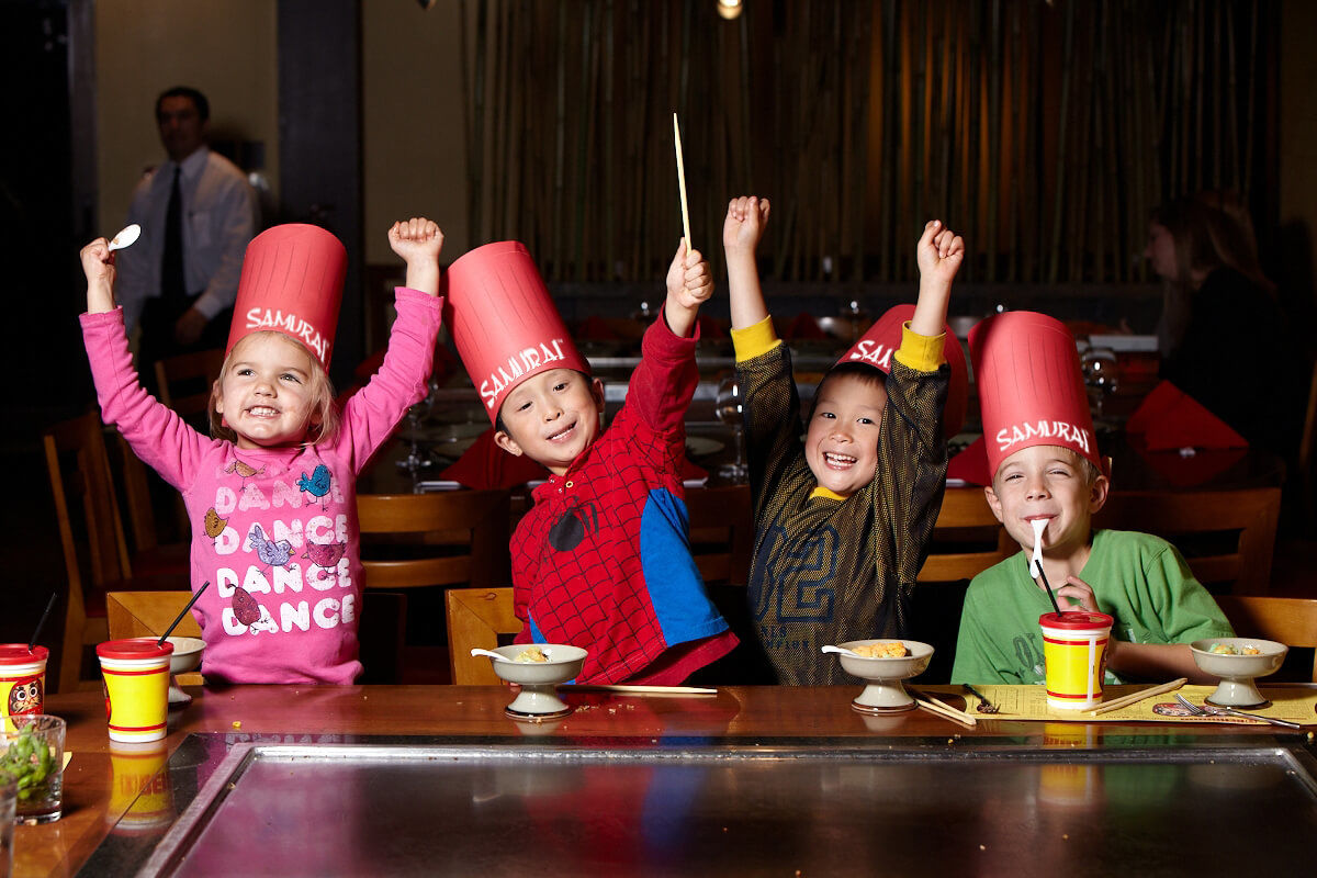 Four children wearing tall red chef hats sit at a teppanyaki grill table, raising their arms. In front of them are bowls of hibachi fried rice and drinks in collectible Samurai mugs.