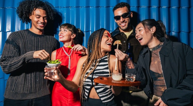 A group of happy young people stood in front of a blue wall eating with chopsticks.
