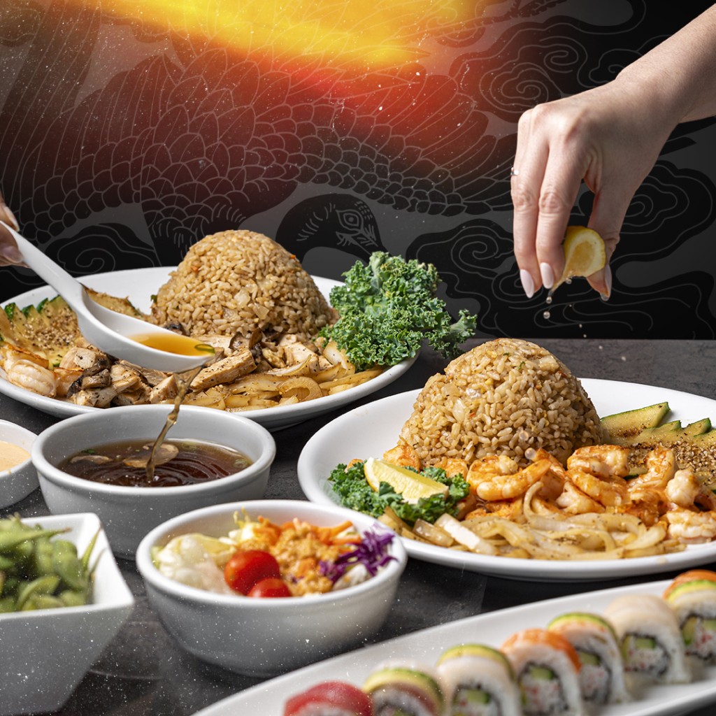 A table set with plates of hibachi fried rice, grilled shrimp, sautéed vegetables, dipping sauces, and a platter of assorted sushi rolls. A hand squeezes a lemon wedge over a plate.