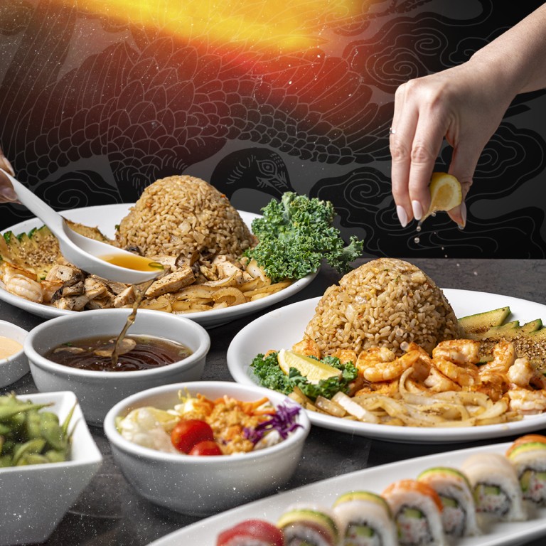 A table set with plates of hibachi fried rice, grilled shrimp, sautéed vegetables, dipping sauces, and a platter of assorted sushi rolls. A hand squeezes a lemon wedge over a plate.