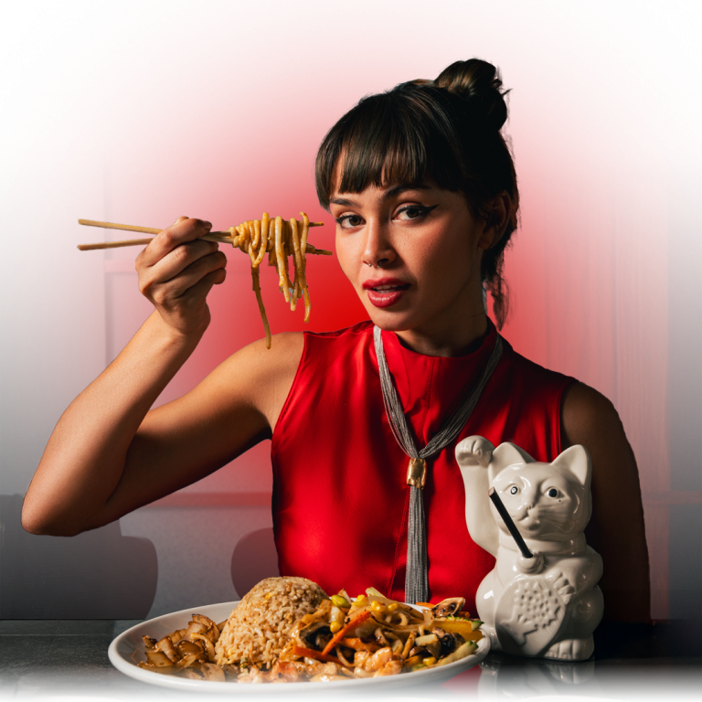 A woman in a red dress holds chopsticks with noodles above a plate of hibachi fried rice, sautéed vegetables, and noodles, next to a white ceramic cat mug with a straw. Gradient circle effect added to background.