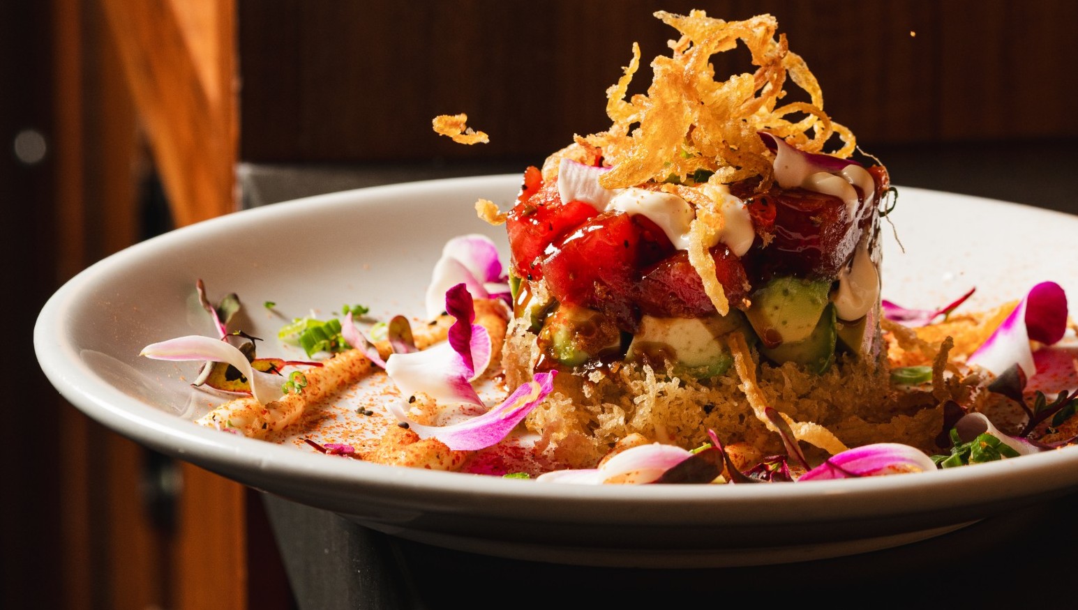 A close up of a white dish with a stack of spicy tuna and avocado on crispy rice, topped with fried onions and garnished with edible flower petals. Blurred background.