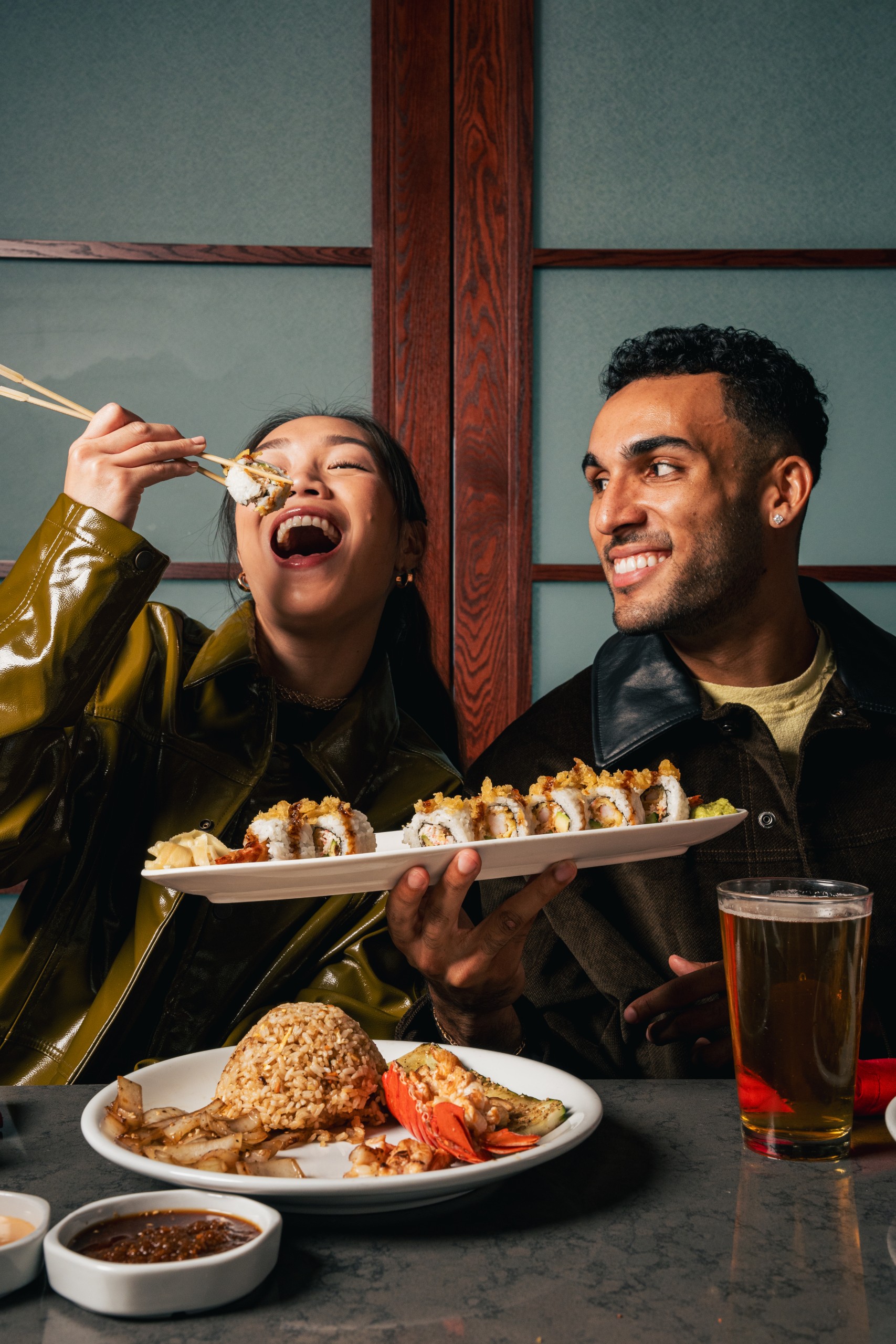 Two people dining at a Samurai restaurant. One holds a plate of assorted sushi rolls; the table features hibachi fried rice, grilled lobster tail, vegetables, dipping sauces, and a glass of beer.