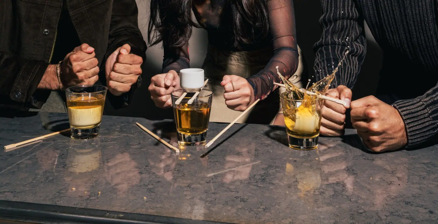 Three people are preparing to do a sake bomb at a bar. Each person has a glass of beer with a pair of chopsticks resting on top, supporting a small cup of sake. The person on the right has just slammed the table, causing the sake cup to drop into the beer and splash.