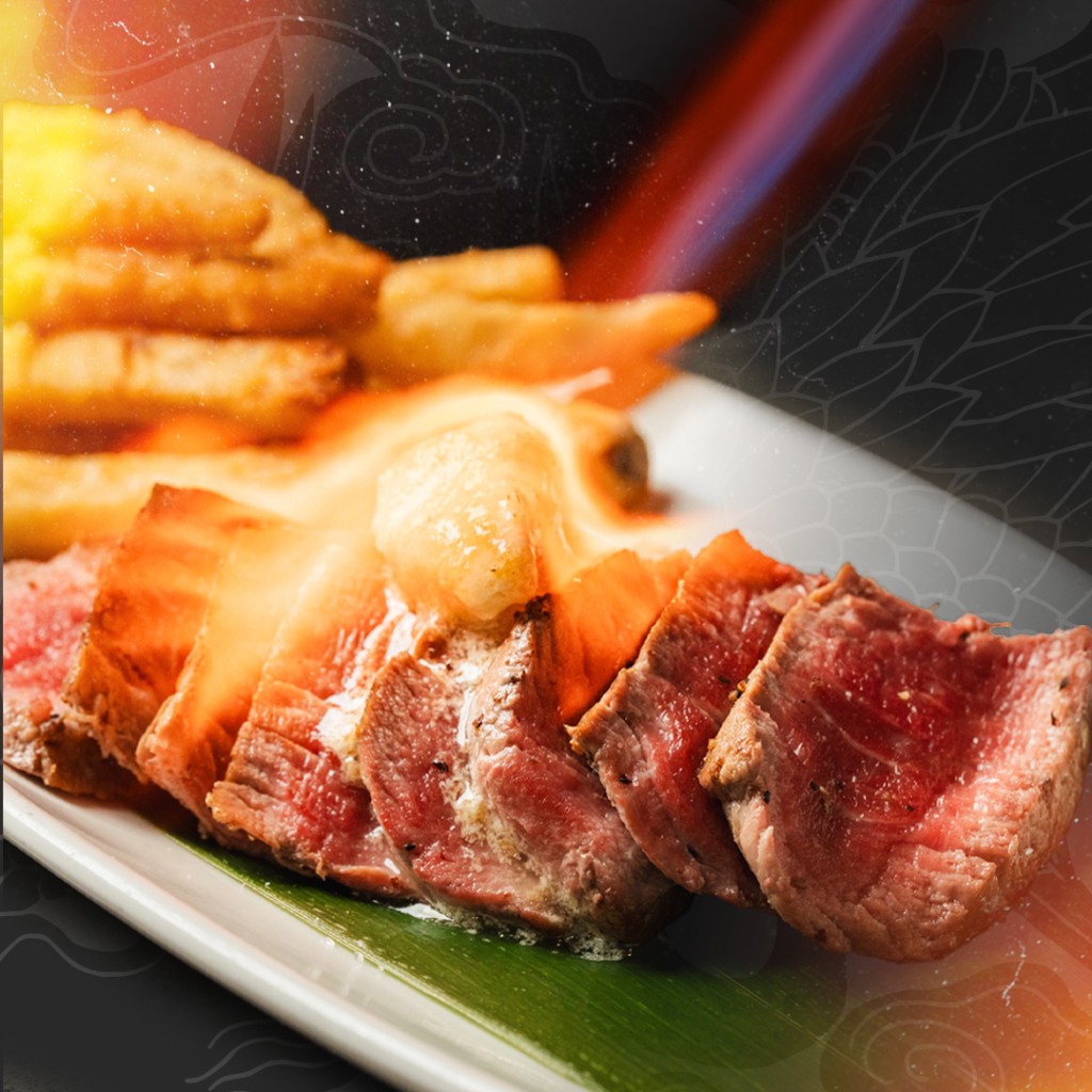 Slices of rare, seared steak topped with melting butter, served on a green leaf alongside a pile of golden fries, with a dramatic flame torching effect in the background.