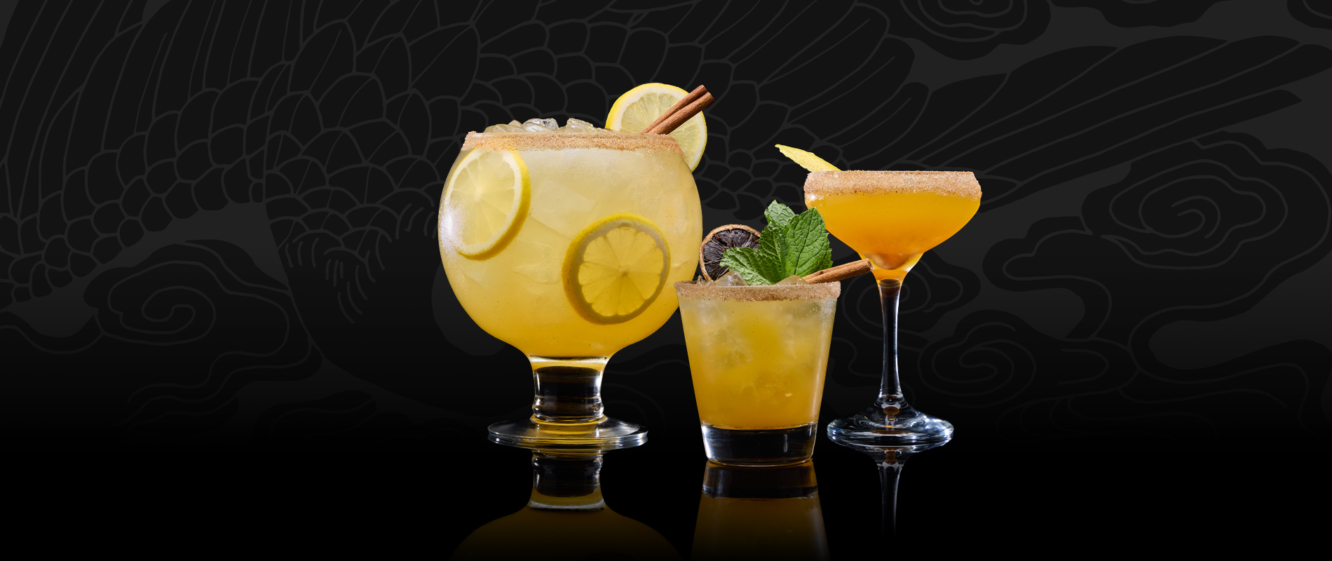 Giant goblet, cocktail glass, and rocks glass filled with golden citrus cocktails, garnished with lemon slices, cinnamon sticks, mint, and sugar rims, set against a black patterned background.