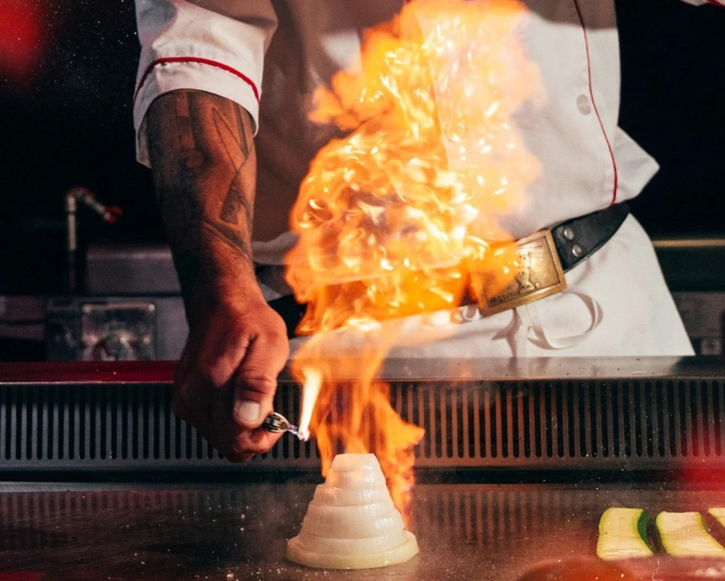 A close up of a hibachi chef igniting a dramatic onion volcano flame on a teppanyaki grill. Focus is on the chefs hand and the flame.
